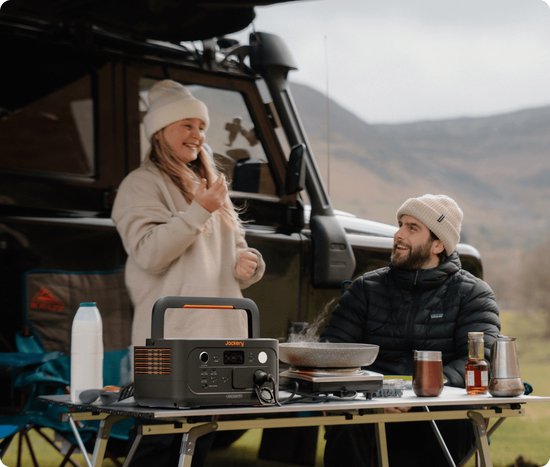 Jackery Explorer 500 powerstation op een campingtafel met een man en vrouw op de achtergrond.