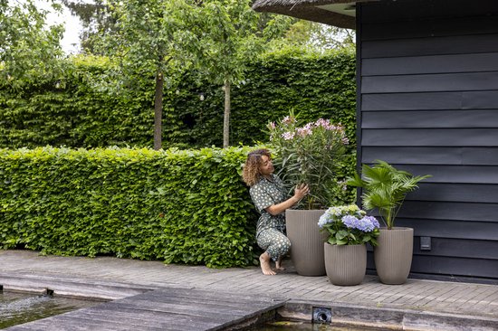 Vrouw in groen jurkje met witte bloemen verzorgt planten in taupe bloempotten naast een houten schuur.