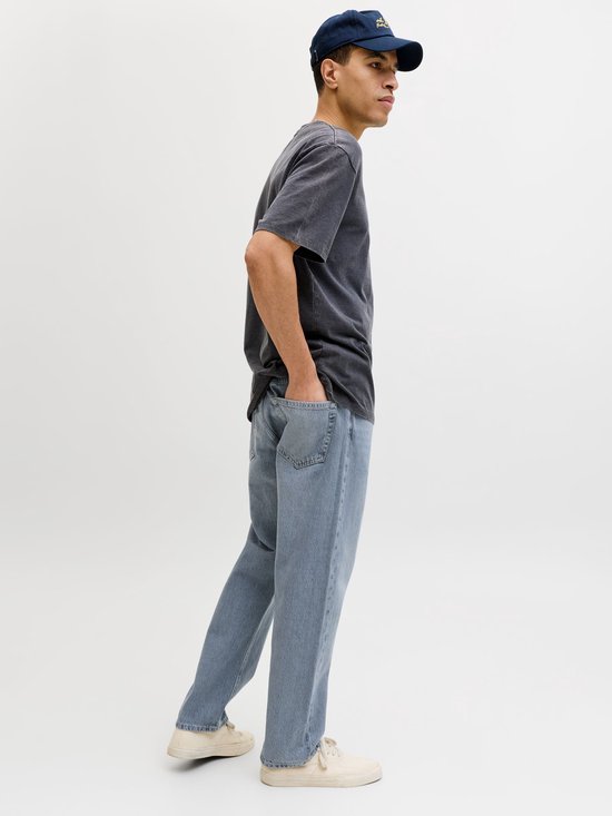 Man met donkergrijs t-shirt, blauwe jeans en pet, staand met handen in de zakken.