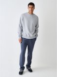Man in een grijze sweater met ronde hals, donkerblauwe joggingbroek en zwarte sneakers.