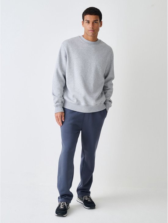 Man in een grijze sweater met ronde hals, donkerblauwe joggingbroek en zwarte sneakers.