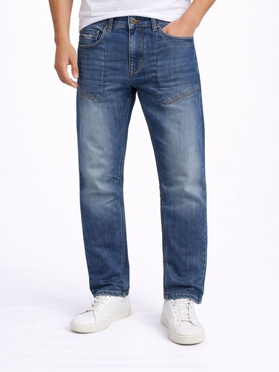 Man in een wit T-shirt en een stonewashed blauwe jeans. De jeans heeft extra naden op de bovenbenen en witte sneakers.
