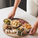 Vrouw met witte broek houdt een houten plank vast met olijven, noten, kaasblokjes, nougat, pesto, grissini en plakjes worst.