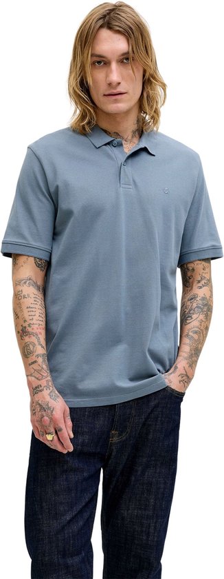 Man met lang blond haar draagt blauwgrijze polo en spijkerbroek, met handen in zijn zakken. Volledige body shot.