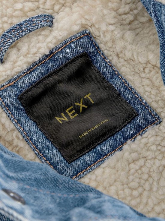 Binnenkant van een blauw denim spijkerjack met crème borgvoering en een zwart "NEXT" label.