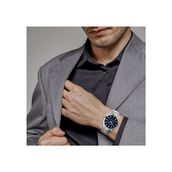 Herenhorloge van Jacques Lemans met blauwe wijzerplaat en zilverkleurige stalen band, gedragen bij een grijs colbert.