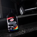Fles Turtle Wax Color Magic Jet Black Wax op een donkere, nat uitziende ondergrond naast een glimmende zwarte auto.