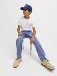 Jongen zittend op een krukje, kijkend naar links, in lichtblauwe spijkerbroek en wit T-shirt.