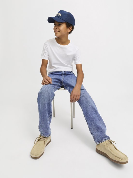 Jongen zittend op een krukje, kijkend naar links, in lichtblauwe spijkerbroek en wit T-shirt.