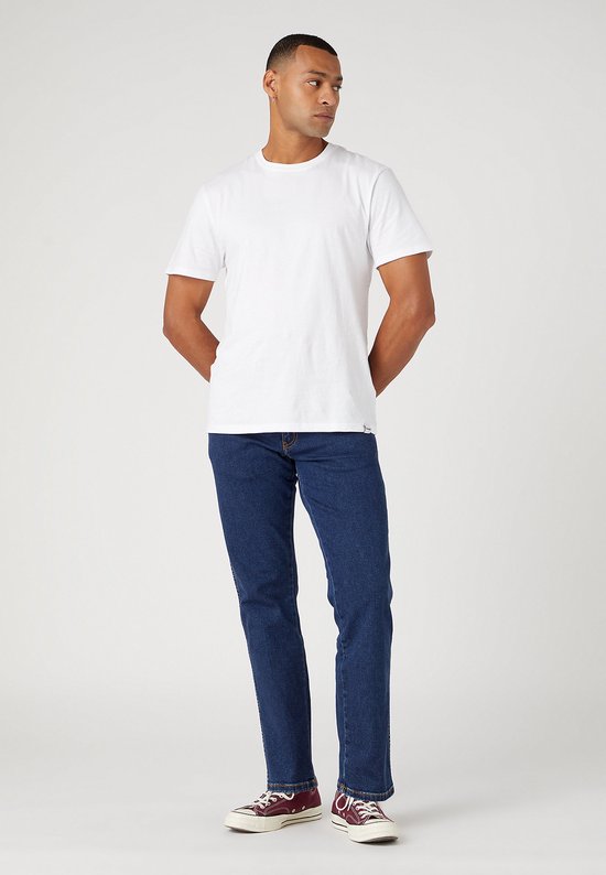 Man in een witte T-shirt en donkerblauwe Wrangler jeans, gedragen met bordeauxrode sneakers.