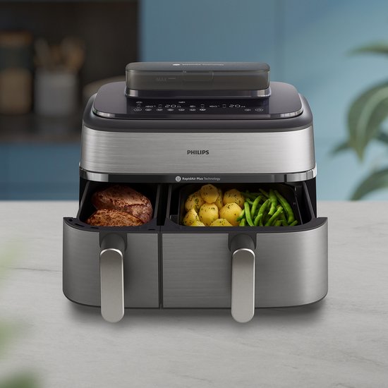 Zilverkleurige en zwarte Philips dubbele airfryer met digitale bediening en twee open lades met kip, aardappelen en sperziebonen, op een aanrecht.
