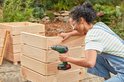 Vrouw met veiligheidsbril boort met een groene Bosch accuboorschroevendraaier in een houten plantenbak.