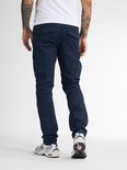 Man in donkerblauwe cargobroek en witte t-shirt, kijkend naar achteren.