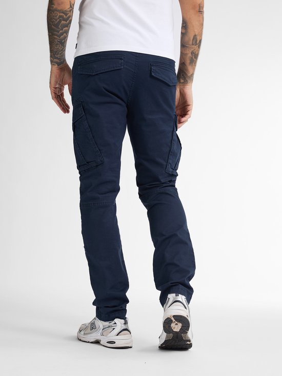 Man in donkerblauwe cargobroek en witte t-shirt, kijkend naar achteren.