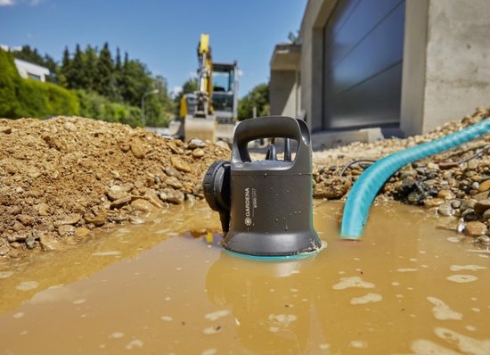 Zwarte en turquoise dompelpomp in een modderige plas, met een blauwe slang en bouwmachines op de achtergrond.