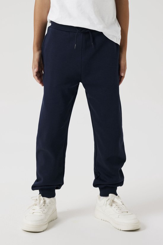 Onderlichaam van een jongen in een donkerblauwe joggingbroek met trekkoord en witte sneakers.