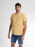 Man met licht getinte huid draagt een donkergeel T-shirt met een klein rond logo met 'P' op de linkerborst en blauwe spijkerbroek.