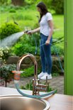 Een vrouw met witte t-shirt en spijkerbroek sproeit met een tuinslang in de tuin, de slang is aangesloten op een kraan boven een spoelbak.