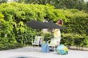 Een vrouw in een gele jurk stelt een grote, donkergrijze Madison zweefparasol op naast witte tuinstoelen.