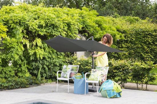Een vrouw in een gele jurk stelt een grote, donkergrijze Madison zweefparasol op naast witte tuinstoelen.