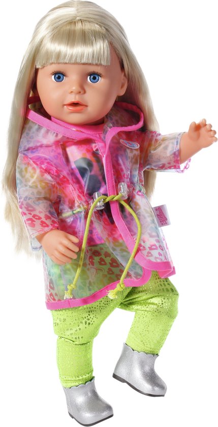 Meisje met blond haar draagt roze shirt met 'BE WILD' tekst, groene broek, zilveren laarsjes en een doorzichtige regenjas met regenboogprint.