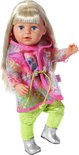 Meisje met blond haar draagt roze shirt met 'BE WILD' tekst, groene broek, zilveren laarsjes en een doorzichtige regenjas met regenboogprint.