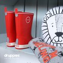 Rode rubberen laarzen met witte rand en logo. Naast een stoffen leeuwenkussen en grijze kleding.