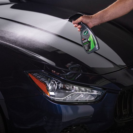 Main vaporisant le flacon gris aux accents verts et noirs sur le capot d'une voiture foncée.