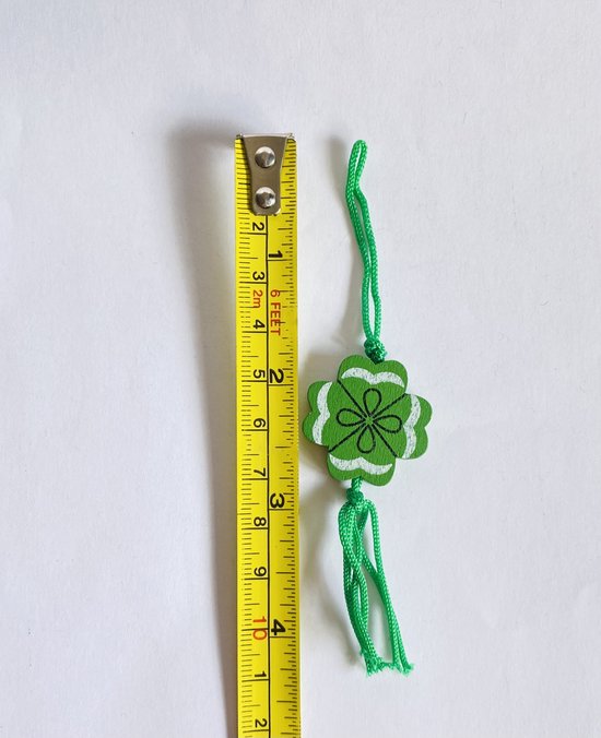 Groen klavertje vier hanger van hout met witte rand en zwart patroon, met koordjes, naast een meetlint.
