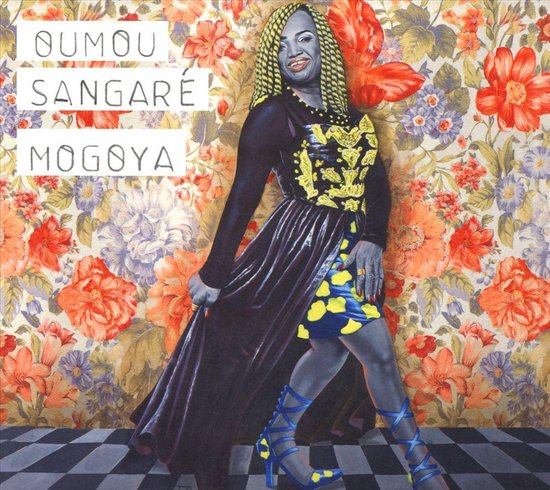 Vrouw in zwart met gele versieringen en een felgekleurde jurk, dansend voor een bloemenbehang. Tekst: OUMOU SANGARÉ MOGOYÁ.