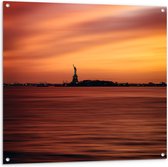 Tuinposter – Vrijheidsbeeld in de Verte met Oranje Lucht - 100x100cm Foto op Tuinposter  (wanddecoratie voor buiten en binnen)