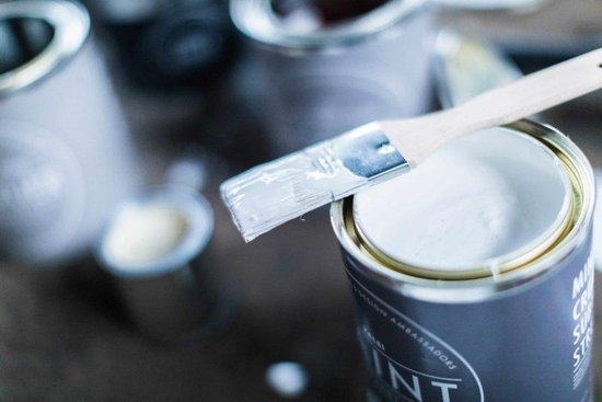 Een kwast met lichte verf erop rustend op een blik met de tekst 'L'Authentique PAINT' en 'Betonlook'.