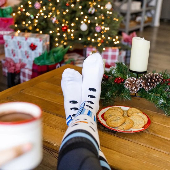 Witte kerstsokken met sneeuwpop-opdruk en blauwe streepjes, gedragen door iemand die aan een tafel zit met warme chocolademelk en koekjes.