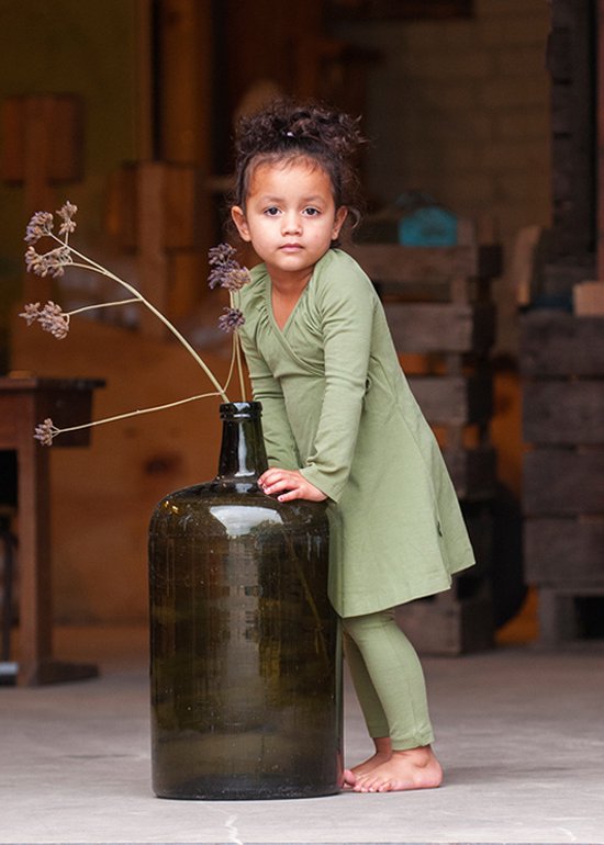 Meisje met krullend haar in lichtgroenwikkeljurkje en legging, staand naast een grote groene fles.