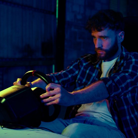 Man met baard speelt een racespel op een Thrustmaster racestuur, verlicht door blauw en groen licht.