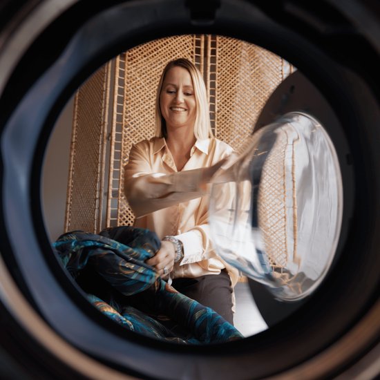 Vrolijke vrouw met blond haar, steekt een bontgekleurd dekbed in een wasmachine.