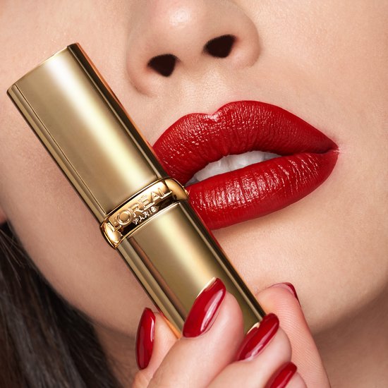 Een vrouw met volle, rode lippen en rode nagels houdt een gouden L'Oréal lipstick vast. De lippenstiftkleur is diep rood.