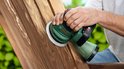 Handen schuren een houten tuinmeubel met een groene Bosch excenterschuurmachine.