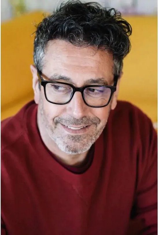 Homme portant des lunettes à monture noire, un pull à col roulé bordeaux, assis avec un sourire.