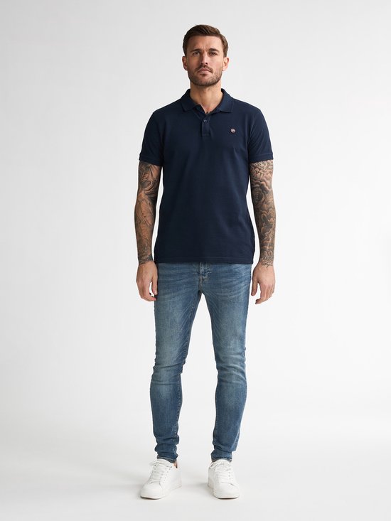 Man met tatoeages draagt een marineblauwe polo en een smalle blauwe jeans.