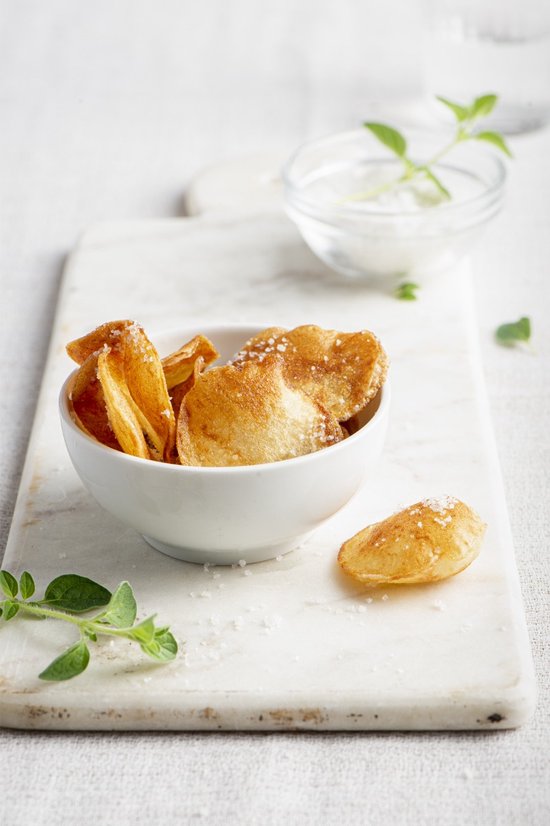 Goudbruine aardappelchips met zout in een witte kom op een marmeren plankje.
