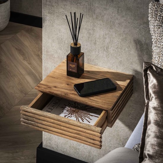 Zwevend nachtkastje van naturel acaciahout met horizontale ribbels en een open lade. Een diffuser en telefoon liggen erop.