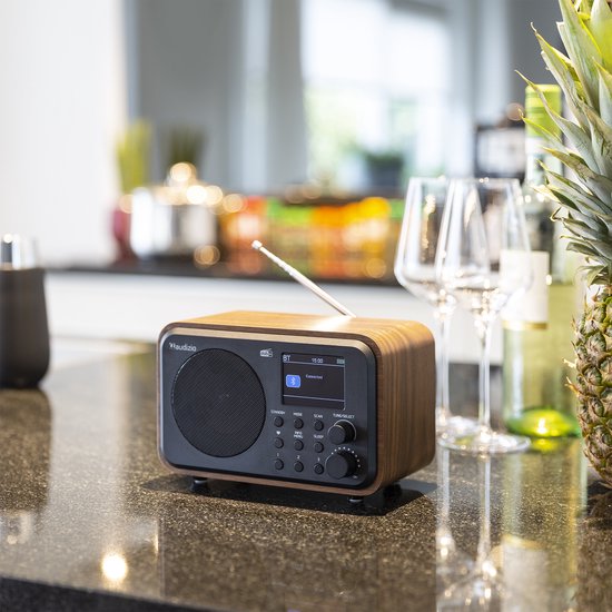 Radio met houten behuizing op een marmeren aanrecht, verbonden met Bluetooth. Naast de radio staan wijnglazen en een ananas.