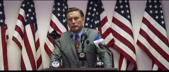 Een man in pak staat achter een lessenaar met microfoons en Amerikaanse vlaggen.
