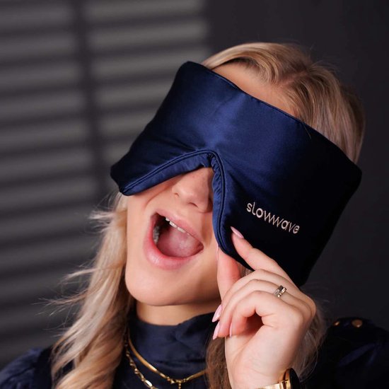 Een vrouw met blond haar met haar mond open, draagt een donkerblauw zijden slaapmasker van Slowwave.