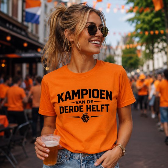 Oranje T-shirt met tekst "KAMPIOEN VAN DE DERDE HELFT" en een leeuwenlogo, gedragen door een vrouw met zonnebril.