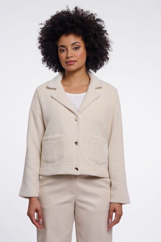 Femme aux cheveux bouclés portant une veste courte couleur crème à col revers, boutons dorés et poches à rabat.