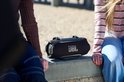 Zwarte JBL Xtreme speaker met schouderband, gefotografeerd tussen twee personen op een betonnen rand.