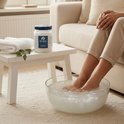 Voetenbad met water en magnesiumvlokken. Een vrouw in beige broek zit ernaast. Pot Zechsal staat op een bijzettafel.