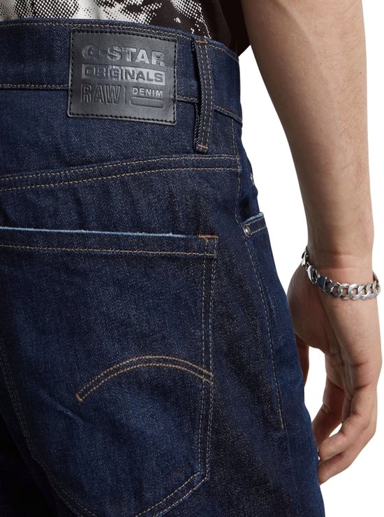 Achterkant van een donkerblauwe G-Star jeans met G-Star Originals label en goudkleurige stiksels. Gedragen door een man met een zilveren armband.
